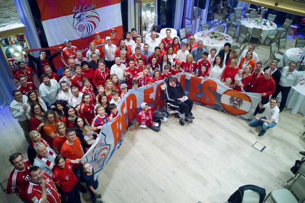 Eine große Gruppe posiert drinnen mit Red Eagles-Fahnen; viele tragen rote Hemden, einige sitzen in Rollstühlen.