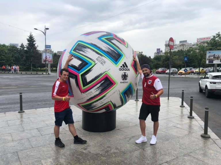 Zwei Männer in Fußballtrikots posieren neben einem riesigen Adidas-Fußball der Euro 2020 auf einer Straße in der Stadt.