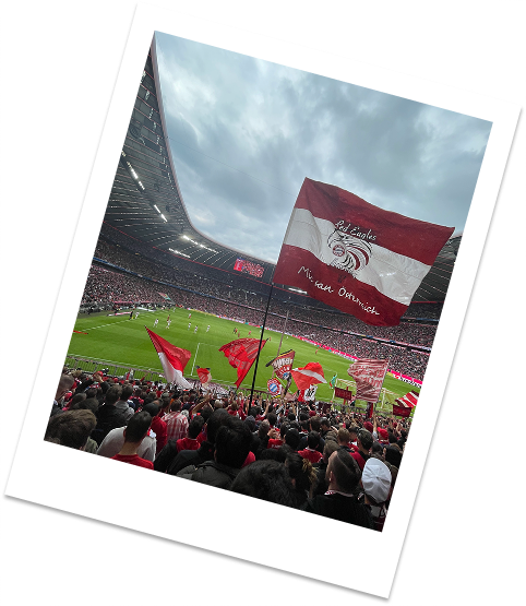 Eine rot-weiße Fahne schwenkende Menschenmenge in einem Fußballstadion während eines Spiels bei bewölktem Himmel.