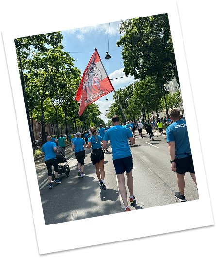 Menschen in blauen Hemden laufen auf einer sonnigen Straße, eine Person hält eine rote Fahne mit einem Motiv.